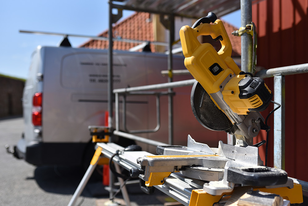 A yellow miter saw is mounted on a stand at a construction site, with a work van parked in the background.