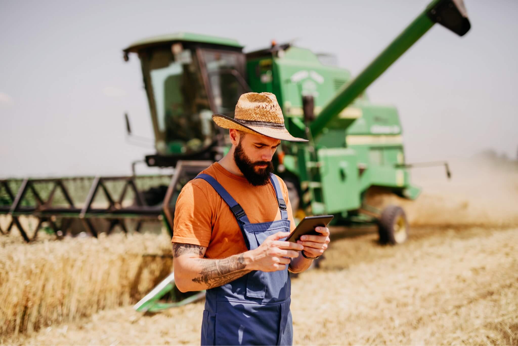 A farmer wearing a hat and overalls stands in a wheat field, checking a tablet with a combine harvester working in the background.