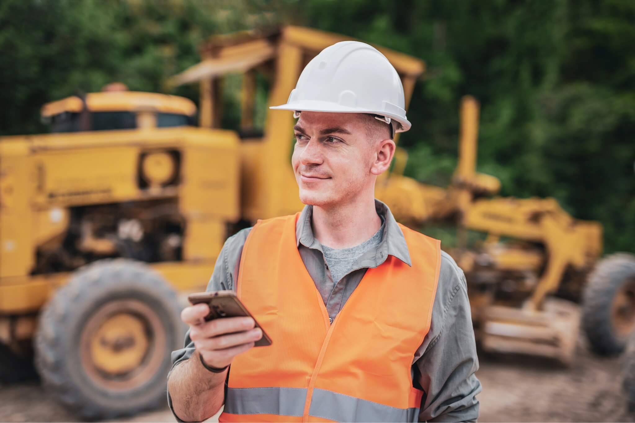 A construction worker wearing a hard hat and safety vest holds a smartphone at a work site with heavy machinery in the background.