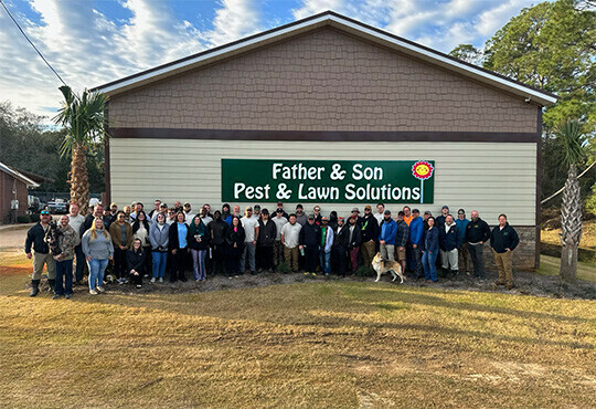 A large group of people stands cheerfully in front of a building with a sign reading "Father & Son Pest & Lawn Solutions," against a partly cloudy sky.