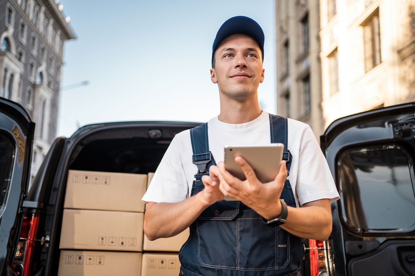 A delivery driver wearing a blue uniform and cap stands in front of a van filled with cardboard packages, holding a tablet.