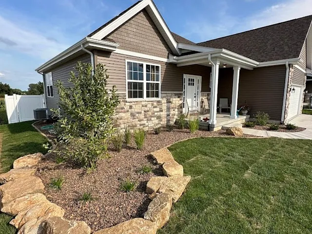 Two workers in neon shirts install landscaping rocks around a house corner. One kneels on a lush green lawn, while the other adjusts rocks near the wall.