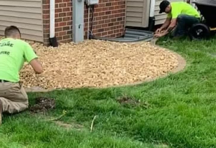 Two workers in neon shirts install landscaping rocks around a house corner. One kneels on a lush green lawn, while the other adjusts rocks near the wall.