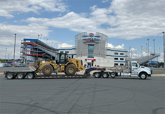 A flatbed truck carries a large yellow bulldozer in front of a modern building with a "zMAX Dragway" sign.