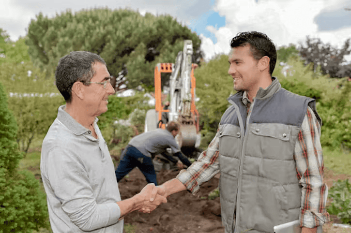 Two men in outdoor clothing shake hands in the foreground, with a worker operating a construction excavator in the background.