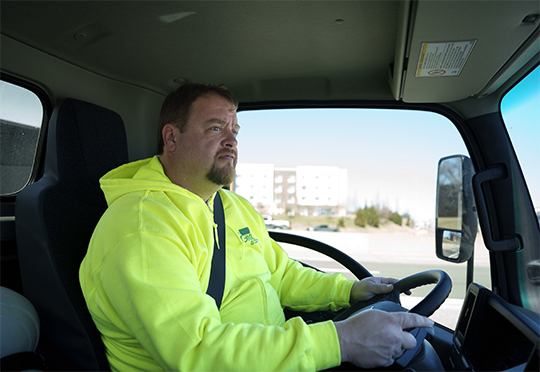 A smiling delivery worker wearing a blue uniform and cap waves to the camera in front of a white work van parked at a residential house.