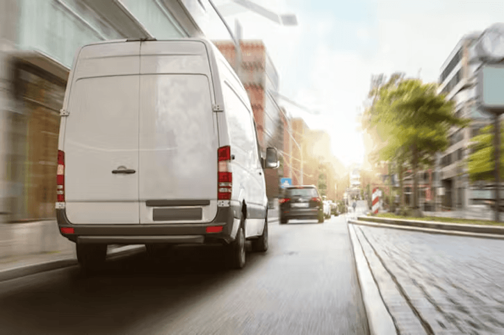 A white delivery van drives down a city street with motion blur, followed by another car in the distance under bright sunlight.