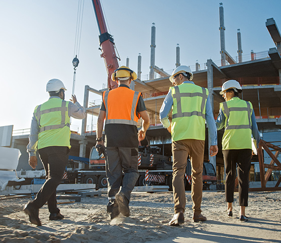 Four construction workers wearing hard hats and high-visibility vests walk away from the camera toward an active building site with a crane.