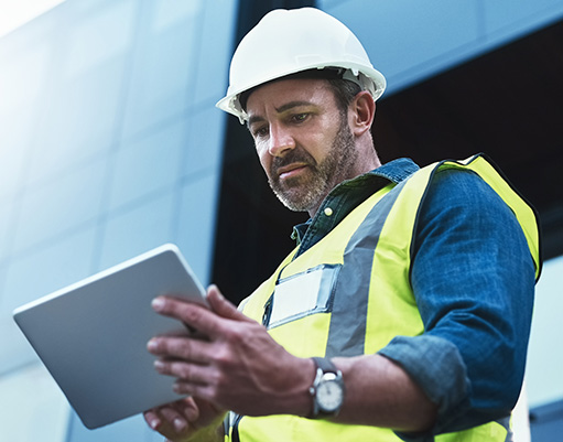 A middle-aged construction professional in a hard hat and high-visibility vest reviews information on a digital tablet outside a modern building.
