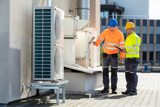 Two technicians in safety gear and hard hats stand on a roof, inspecting HVAC equipment.
