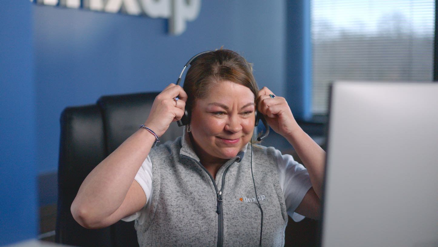 A smiling woman wearing a grey vest and a headset sits at a desk in front of a computer screen.