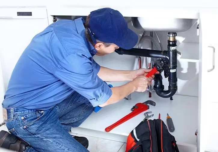 A plumber wearing a blue shirt and cap kneels under a sink to repair the plumbing with a wrench.