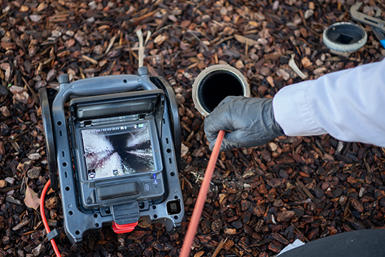 A person wearing a grey glove feeds a camera cable into an open pipe while monitoring the internal view on a portable inspection device placed on mulch.