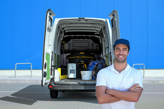 A smiling delivery person with arms crossed stands in front of a white van with its rear doors open, revealing equipment inside against a bright blue wall.