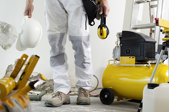 A worker in protective clothing stands in a construction area holding a hard hat and earmuffs, with an air compressor and tools nearby.