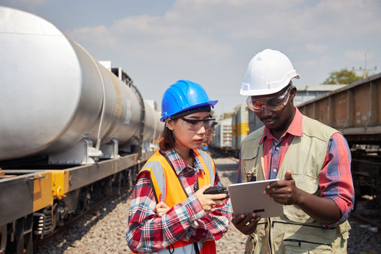 Two railway workers wearing hard hats and safety gear stand next to a train tank car while reviewing information on a tablet.