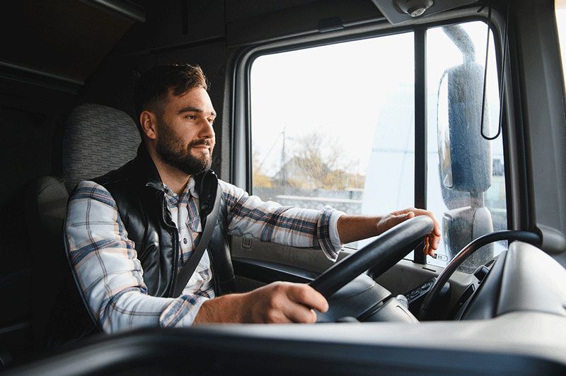 A young man with a beard, wearing a plaid shirt and a black vest, sits in the driver's seat of a truck with his hands on the steering wheel.