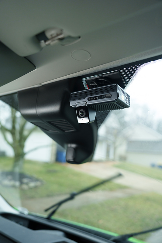 A dashboard camera is mounted on the windshield of a vehicle near the rearview mirror assembly, with a blurred residential view visible through the glass.