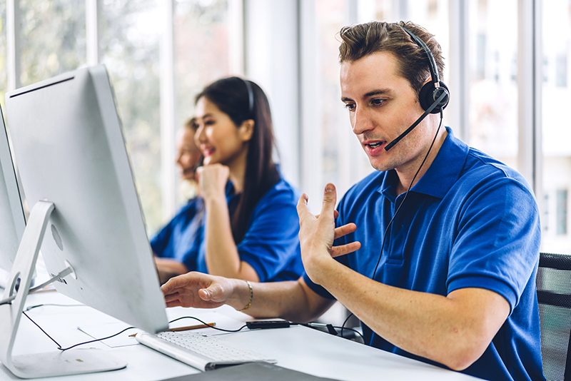A male customer service representative wearing a headset and blue polo shirt talks to a customer while looking at his computer screen in a modern office, with a female colleague working in the background.