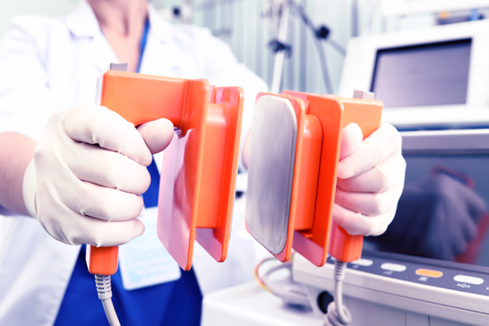A healthcare professional wearing medical gloves holds a pair of orange defibrillator paddles in front of a medical monitor.