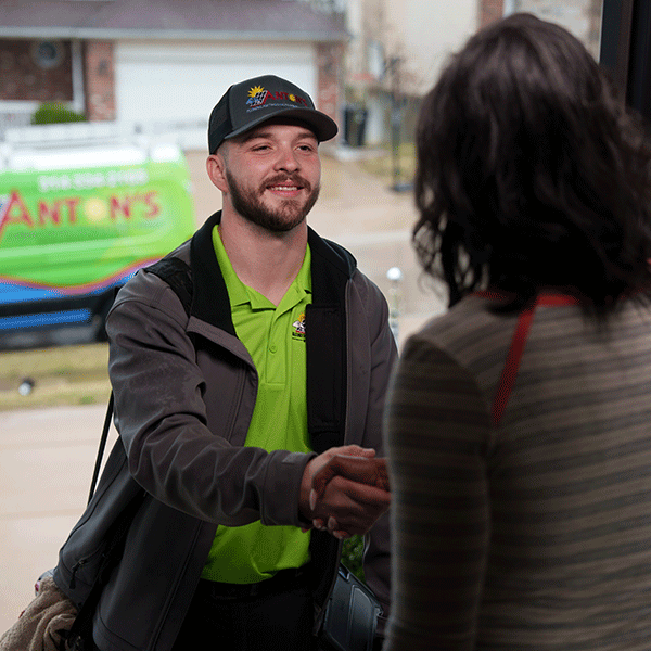 A smiling service technician in a neon green shirt and cap shakes hands with a customer at her front door, with a company van visible in the driveway behind him.
