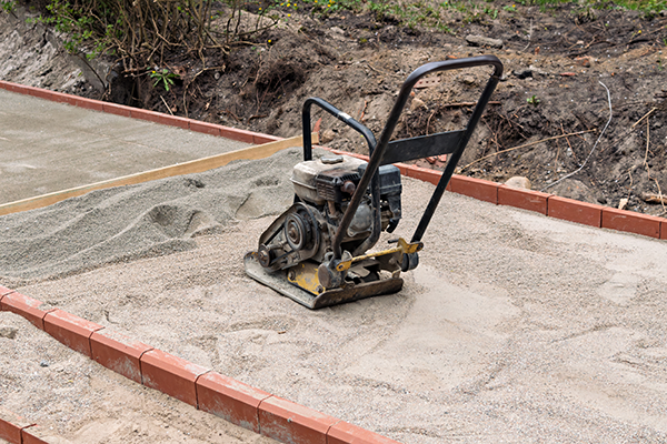 A plate compactor sits on a bed of sand between rows of red bricks at a construction site.