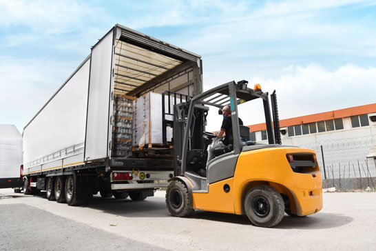 A forklift operator loads palletized goods into the back of a large semi-truck trailer outdoors.